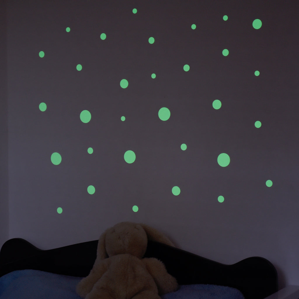 Leuchtpunkte in verschiedenen Größen an einer Wand im Dunkeln, davor Bett mit Plüschhase
