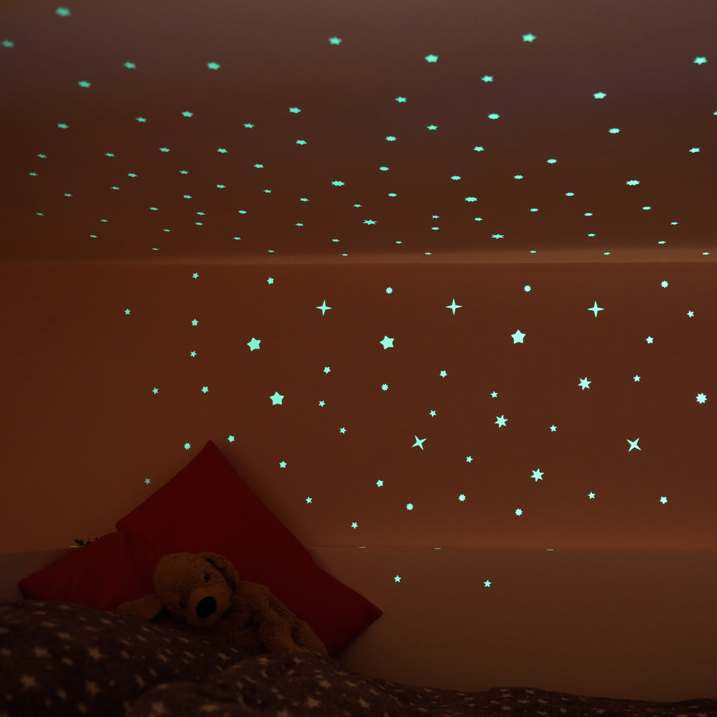 Leuchtsterne an der Zimmerdecke und Wand eines Kinderzimmers im Halbdunkel, Teddy auf dem Bett