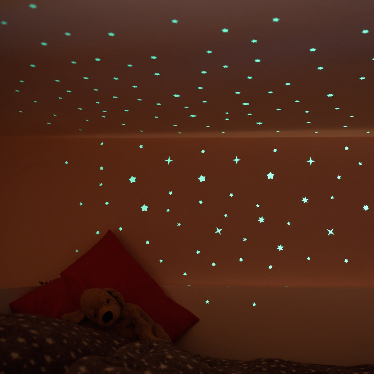 Leuchtsterne an der Zimmerdecke und Wand eines Kinderzimmers im Halbdunkel, Teddy auf dem Bett